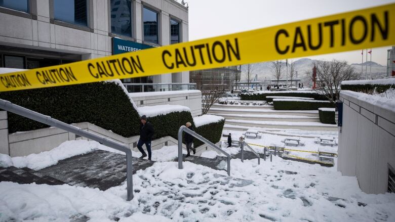 Yellow caution tape runs diagonally in the forefront of the photo, blocking off snow-covered outdoor steps. Two people can be seen walking up cleared steps to the left.