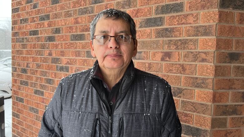 A man with glasses and a jacket stands in front of a brick wall and looks at the camera.