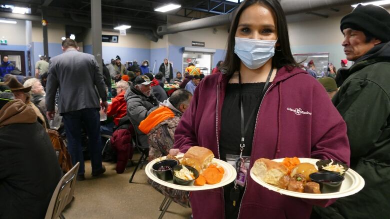 A woman with long dark hair wearing a maroon sweater and a face mask holds two plates of food. She is standing in front of a room full of people.