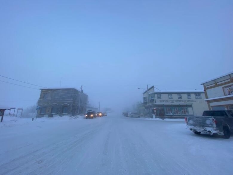 A view up a snowy road, with a couple of buildings and vehicles visible in the distance.