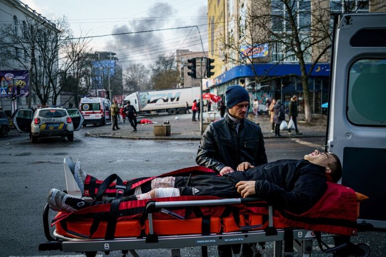 An injured man lies on a stretcher as another man stands over him on a street, while smoke rises in the distance.