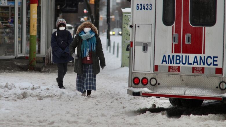 Two women wearing winter coats walk along a snowy sidewalk as an ambulance turns a corner.