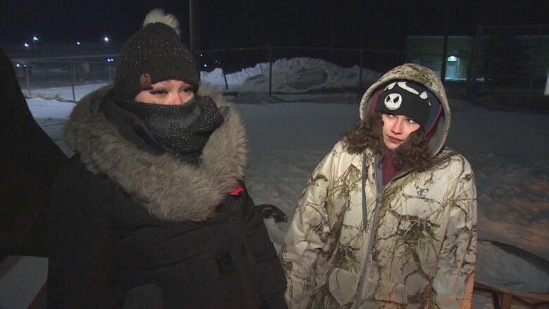 In the snow, a woman in a black coat stands next to a shorter woman in a cream coloured coat.