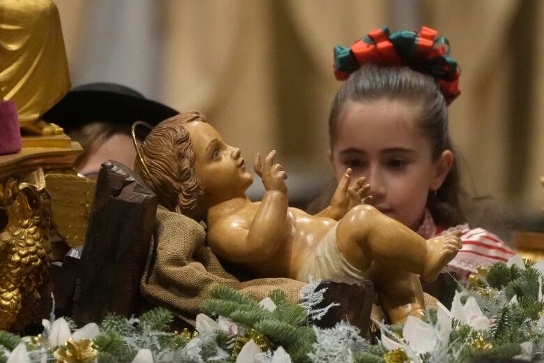 A girl with a large red and green bow in her hair looks at a small statue of a baby. 