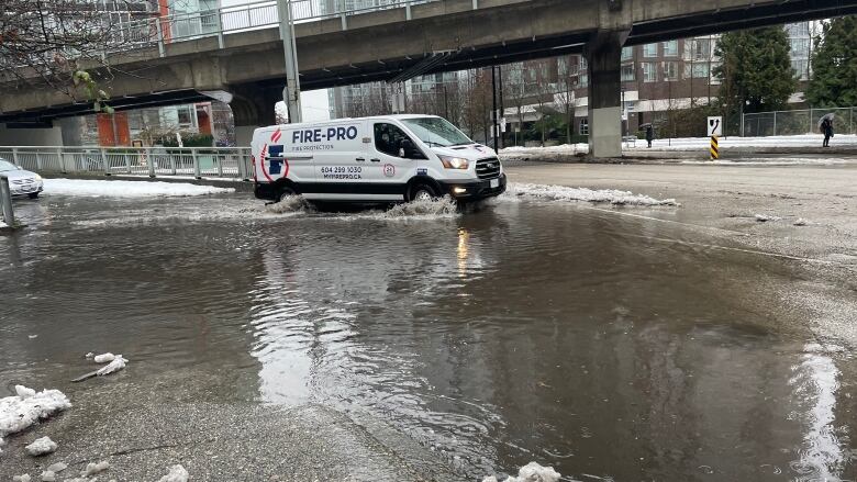 A truck spews up water as it traverses a sludgy water-pooled road, with apartment buildings visible in the background.