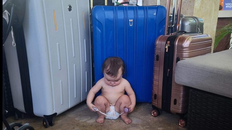 A baby in a diaper sits in front of luggage. 