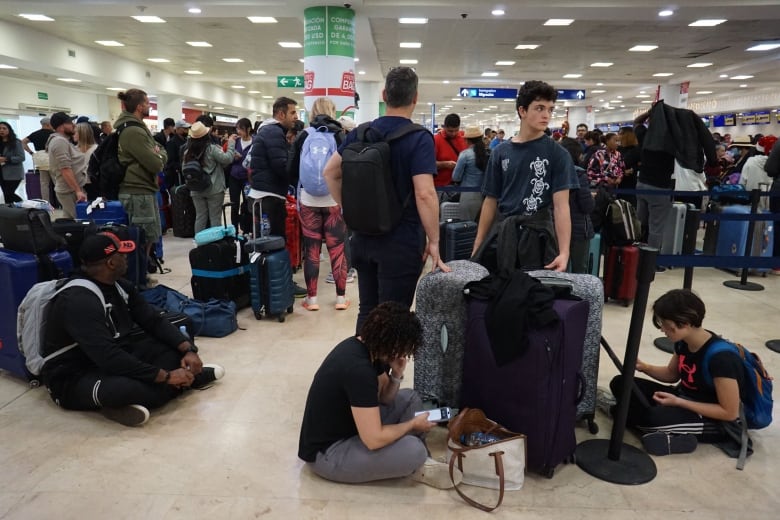 A lot of people sit or stand around luggage in an airport. Some people are sitting on the floor looking at their cellphones.