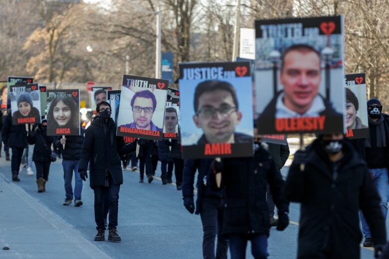 People dressed in black hold signs, each with one person's face and name on them, while walking in a line down a street.