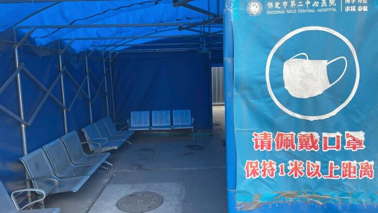 View seen inside a big blue tent that has a sign with Chinese-language characters and a white medical mask on it. 