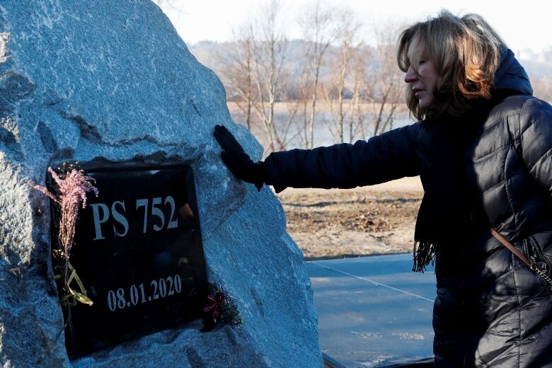 A woman in a black parka stretches her right hand out to touch a large boulder with a plaque on it that reads 