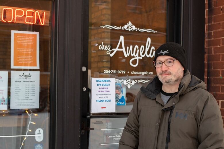 A man wearing a big winter jacket and toque stands outside a door that says 