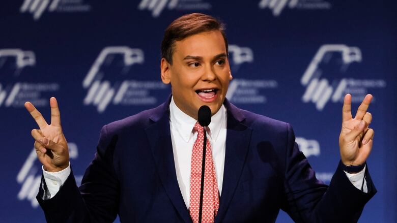 A man in a dark blue suit and red tie speaks into a microphone while raising both his hands and making quotation marks or V signs with his fingers.