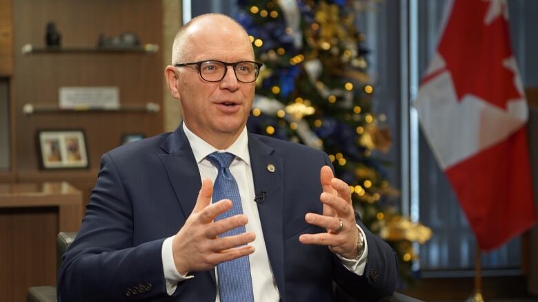 A man wearing a suit is sitting and talking in an office, with a Christmas tree and Canadian flag in the background.