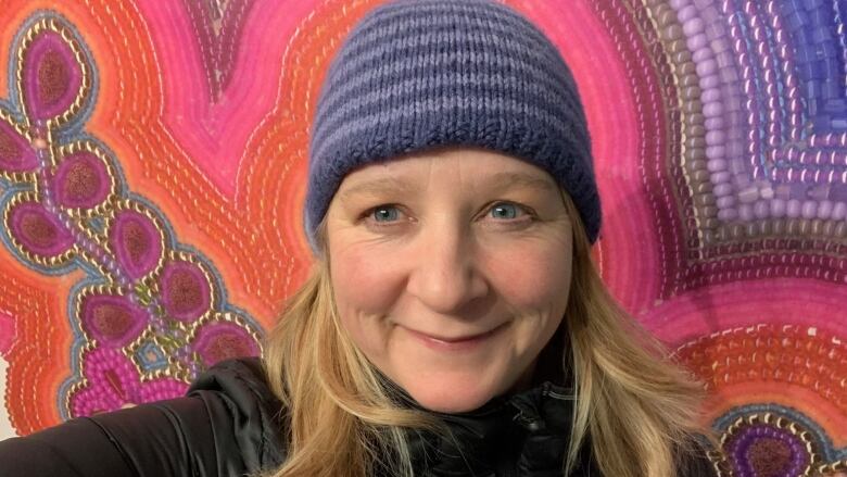 A smiling woman in a toque stands in front of a colourful beaded tapestry.