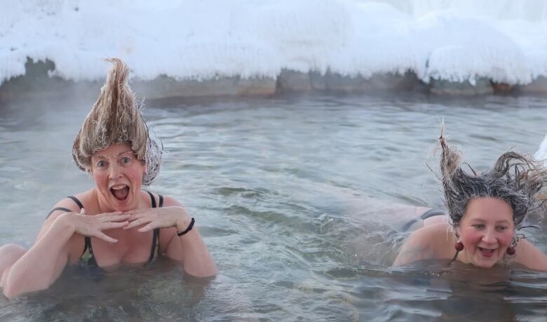 Two women, with their hair standing up frozen, are seen soaking in hot springs on a snowy winter day.