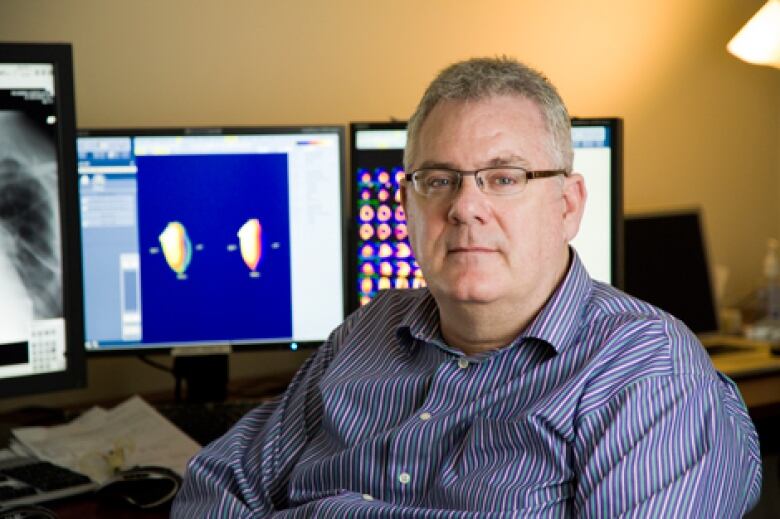 A man wearing glasses and a blue shirt sitting in front of two computer screens. 