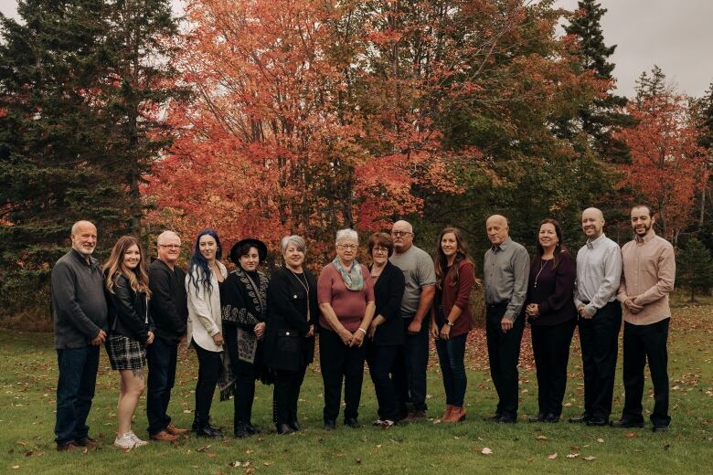 A family of 14 stands in a line posing in front of fall foliage.