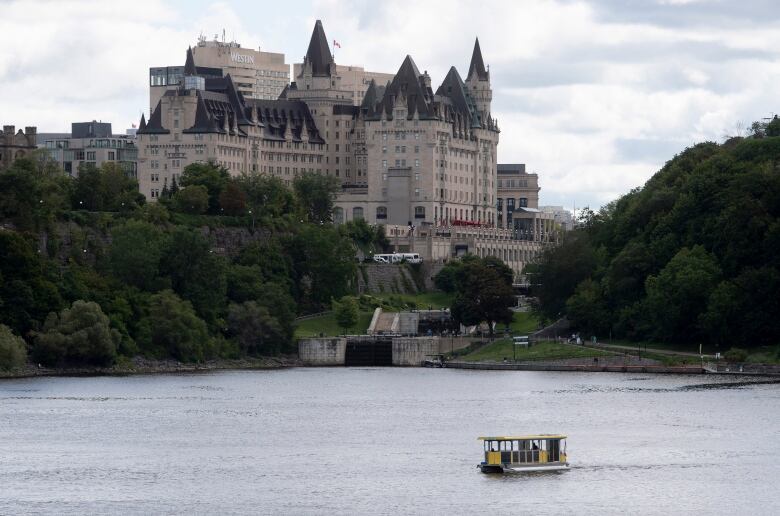 A water taxi crosses a body of water, with trees and a large castle-like building in the background.