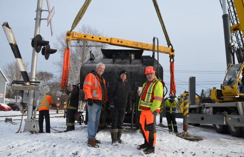 Three people, two in safety jackets, stand in front of a black railway tender car. There is snow on the ground. 