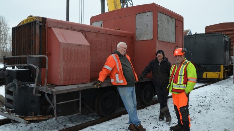 Three volunteers pose on front of a terracotta coloured small locomotive sitting on tracks. There is snow on the ground. 