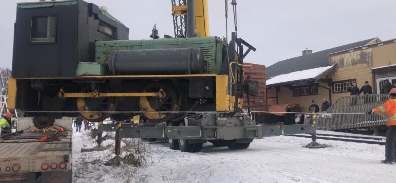 A small green and black locomotive is lowered by a crane to the snow-covered ground.