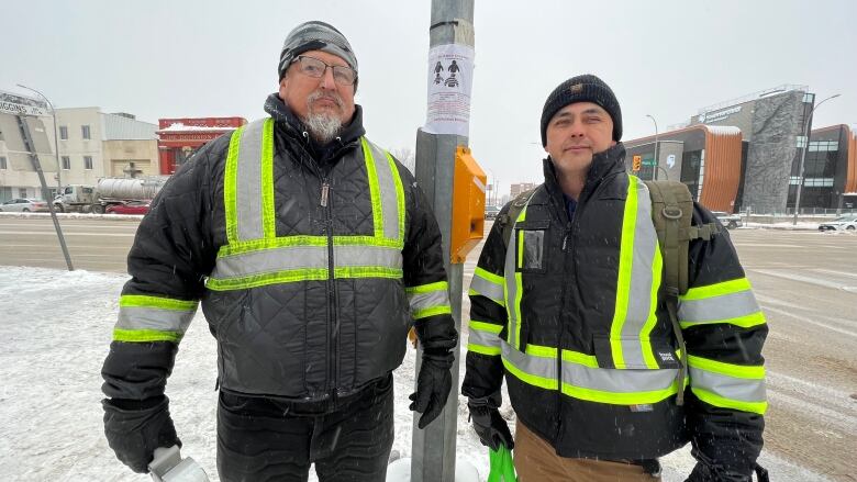 Two men wearing reflective vests stand on a snowy sidewalk on Main Street. A poster is taped to a light pole behind them. 