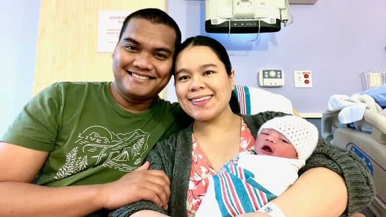 Two smiling parents hold up a crying baby in a hospital room.