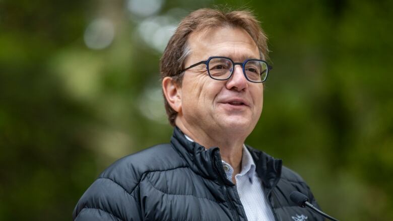 A man in a light jacket and glasses speaks at a microphone. 