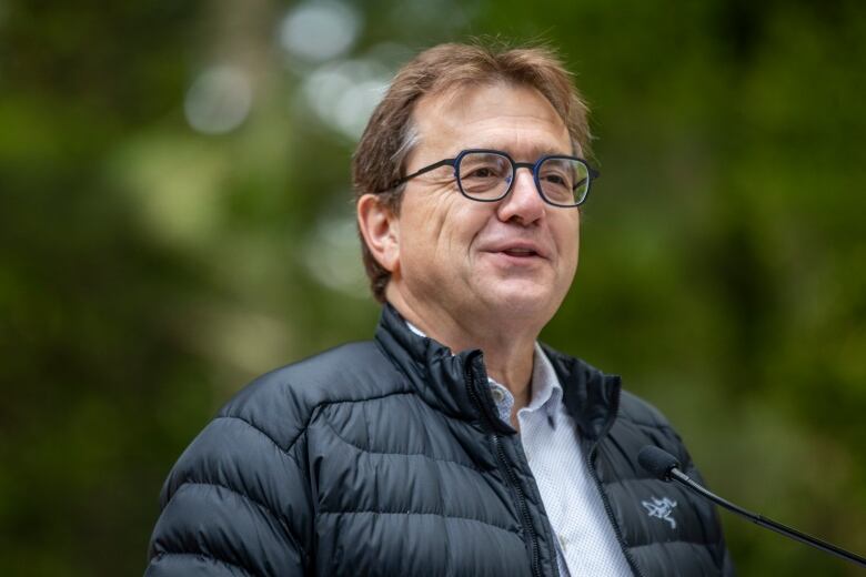 A man in a light jacket and glasses speaks at a microphone. 