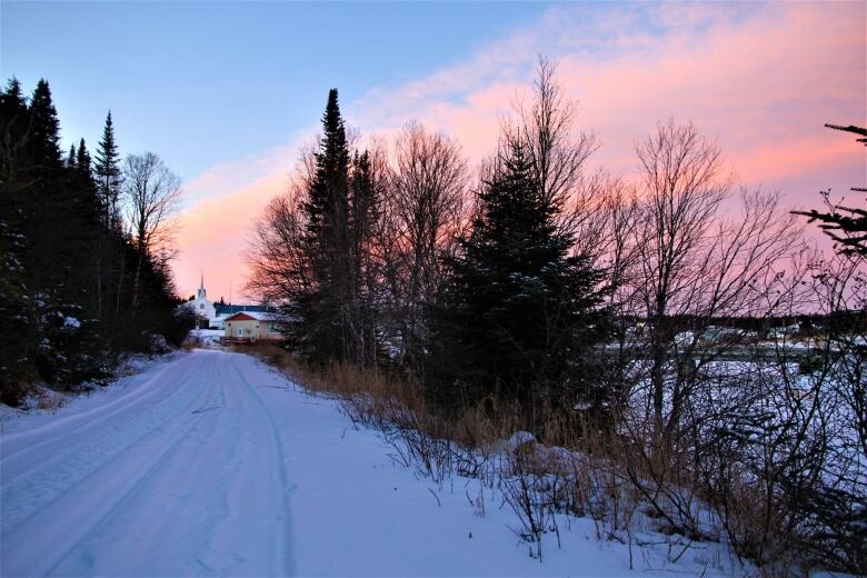 The sun sets, turning the sky shades of pink and purple. A long snow-covered road surrounded by trees leads to a white church in the distance. 