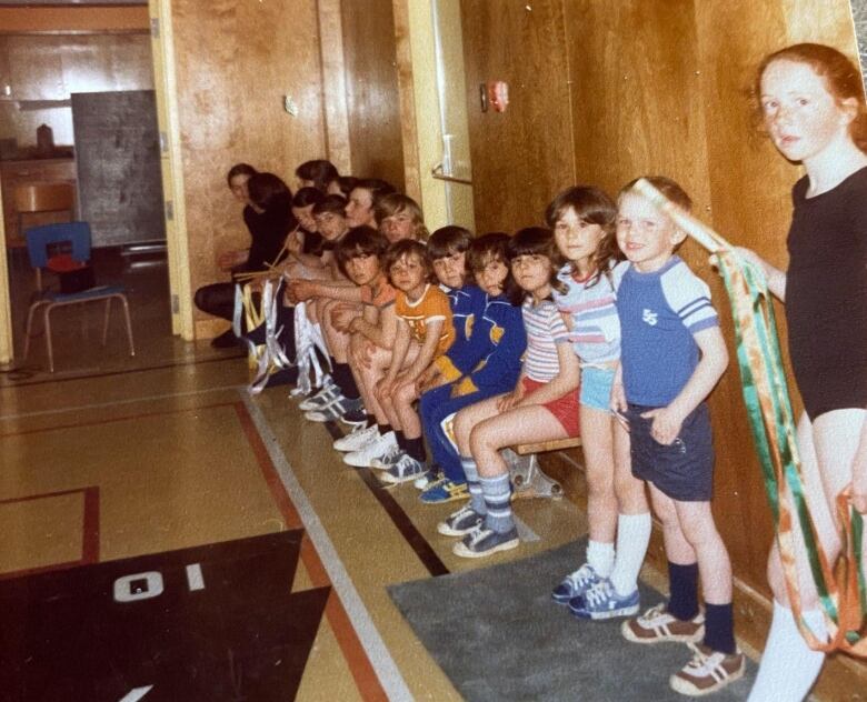 A group of a dozen children sitting on a bench lean in to look at the camera. They are dressed in running shoes and shorts. 