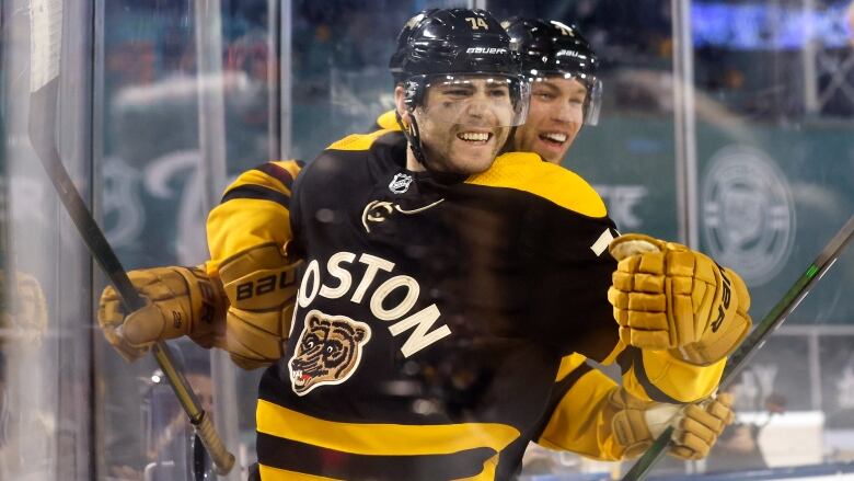 A hockey player wearing black and yellow gear celebrates his goal with a smiling face as he's hugged by a teammate from behind. They are both close to the glass that separates the audience from the rink.