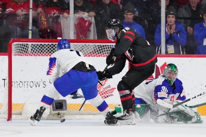 A Canadian hockey player wearing black uniform guides the puck into the net after beating the Slovakian goaltender, seen not able to cover the net in the right side of the picture. A Slovakian defender is also seen in front of the goal but behind the Canadian player.