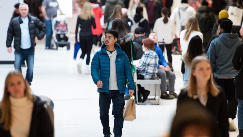 People walk between stores in a mall corridor, some of them holding shopping bags.