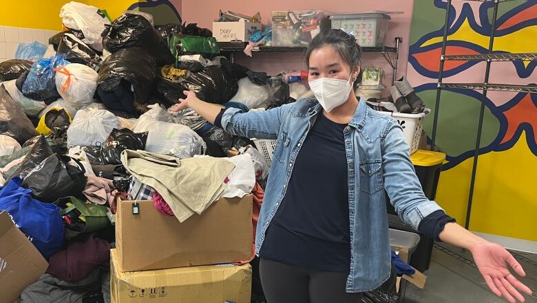 A woman in black pants, black crew neck t-shirt, and light blue button up overshirt stands in front of a pile of donations in bags and bins. The pile is taller than she is, as she holds her arms out to her sides in front of it. 