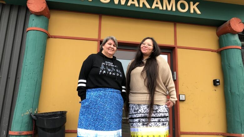 Two women wearing colourful ribbon skirts stand in front of building.