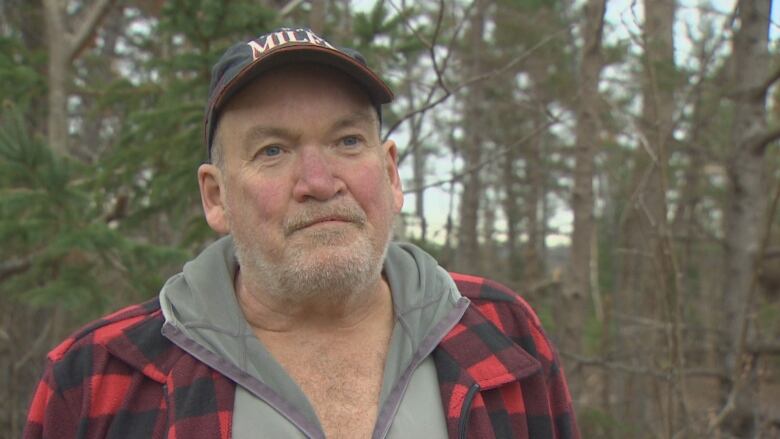 A man stands in a forested area while wearing a baseball cap and a red flannel jacket.