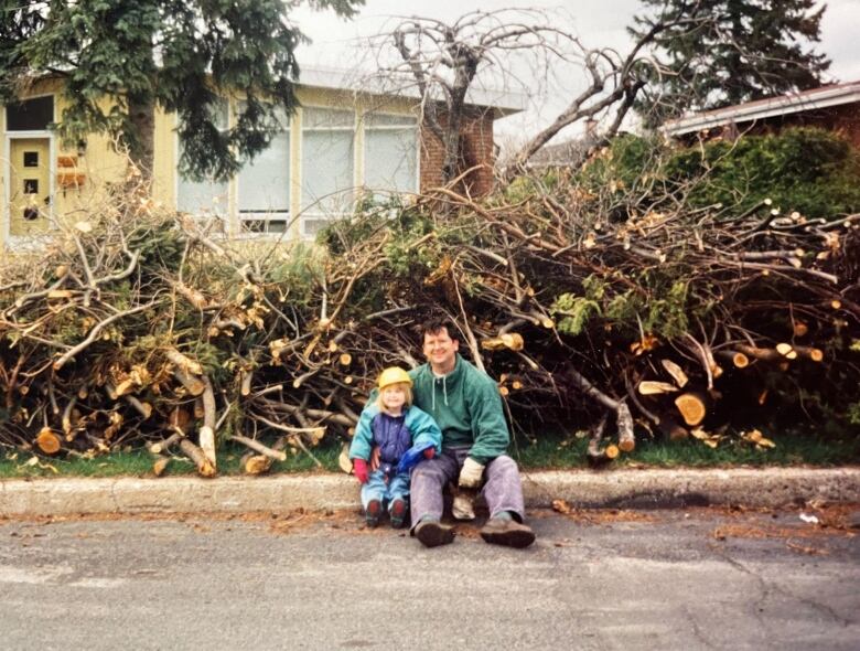 A young child in a plastic hardhat beside a man in a hoodie. In the background a huge pile of broken branches.