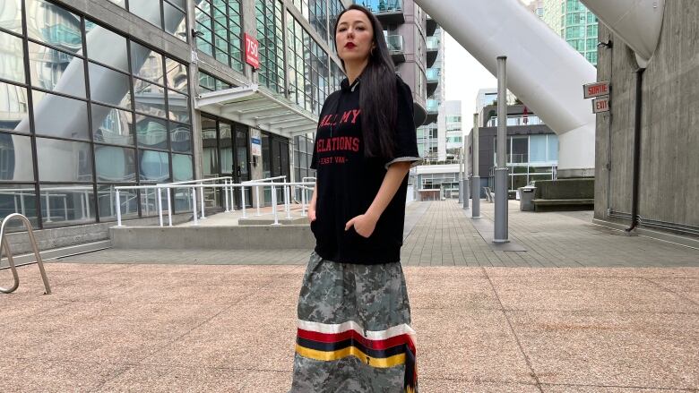 A woman wears an All My Relations basketball shirt with a camo ribbon skirt featuring the words East Van. 