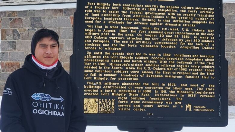 A boy in a hoodie stands beside a historical information sign.