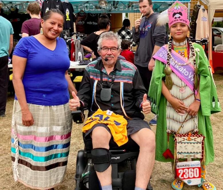 A woman in a ribbon dress, a man in a ribbon skirt and a girl in powwow regalia pose for a photo.