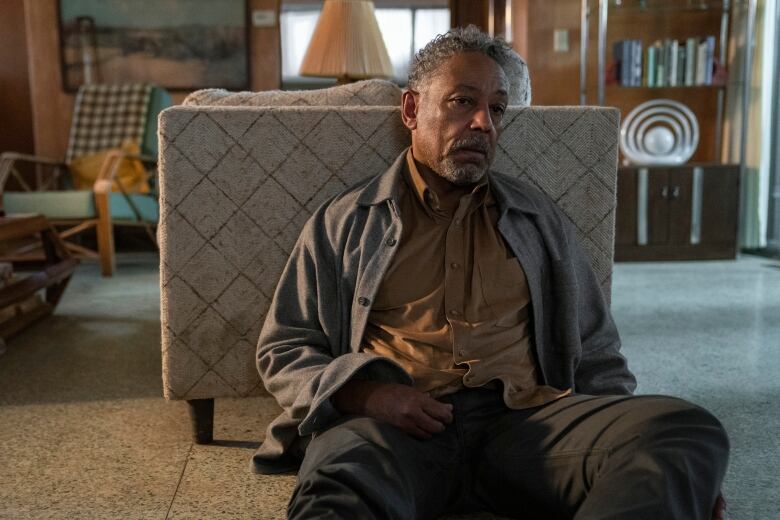 Grey haired man in grey jacket and brown shirt,pants, leaning against sofa flashing a disappointed frown