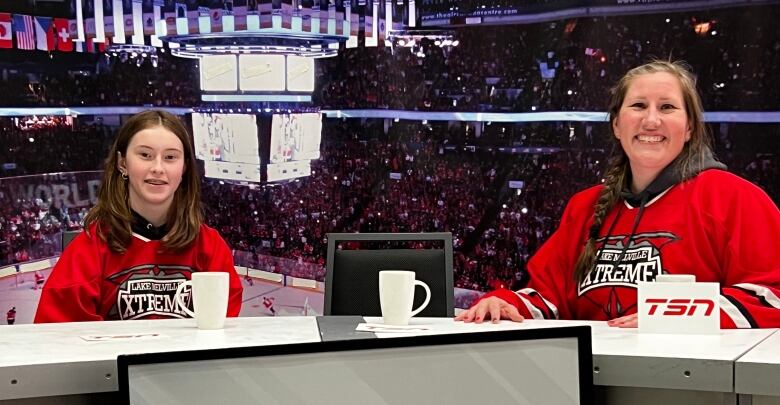 A mother and daughter sit at a table used by TSN for broadcasts in a hockey rink. 