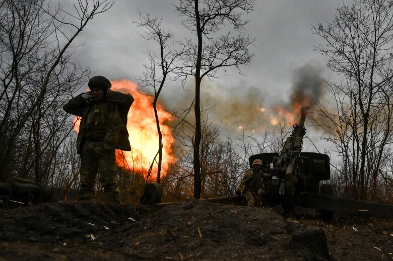 A burst of fire is seen behind a man dressed in combat-style gear. 