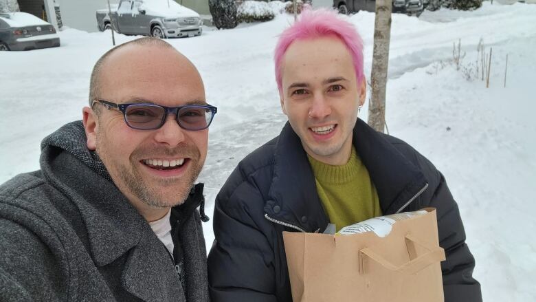 Two men, one bald and the other with pink hair, smile as they stand on a snow-covered street.