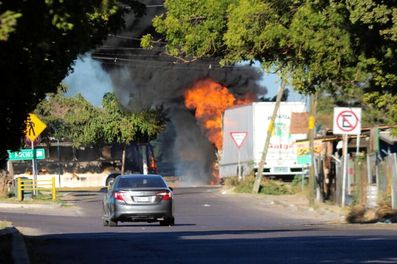 A dark grey car is seen stopped on a road. Several metres away, a vehicle is on fire.