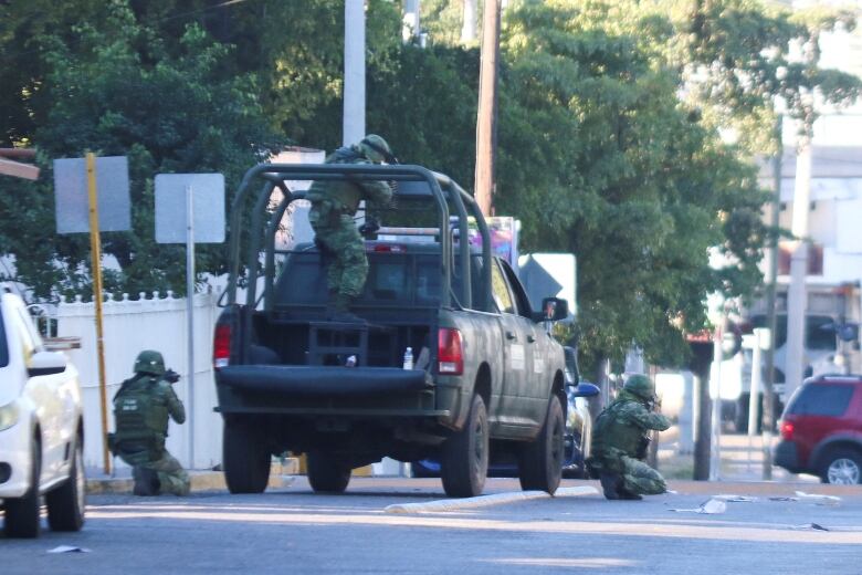 Two soldiers dressed in camouflage and helmets, and pointing guns, are crouched on either side of a pickup truck. A third soldier is standing in the bed of the pickup truck.