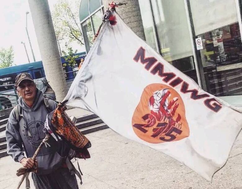 A man stands with a flag. The flag has the letters MMIWG and the number 215. 