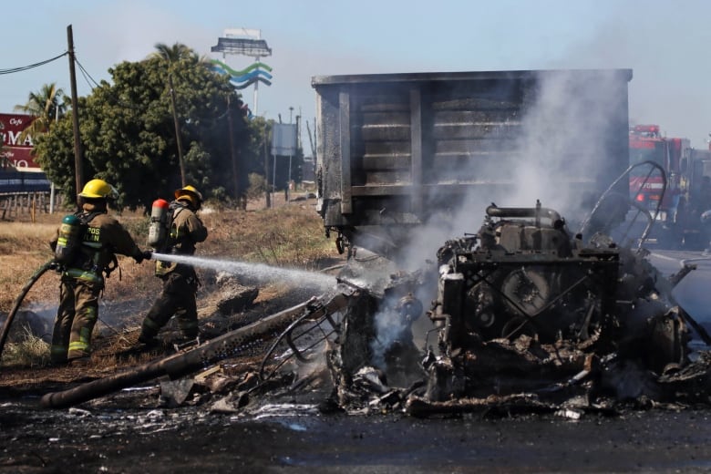 A firefighter points a fire hose at a burned-out vehicle as another firefighter walks in front of them.