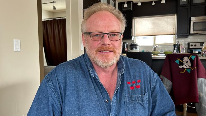 A man smiles for the camera, wearing a blue denim shirt, in his living room. Part of his kitchen in the background.